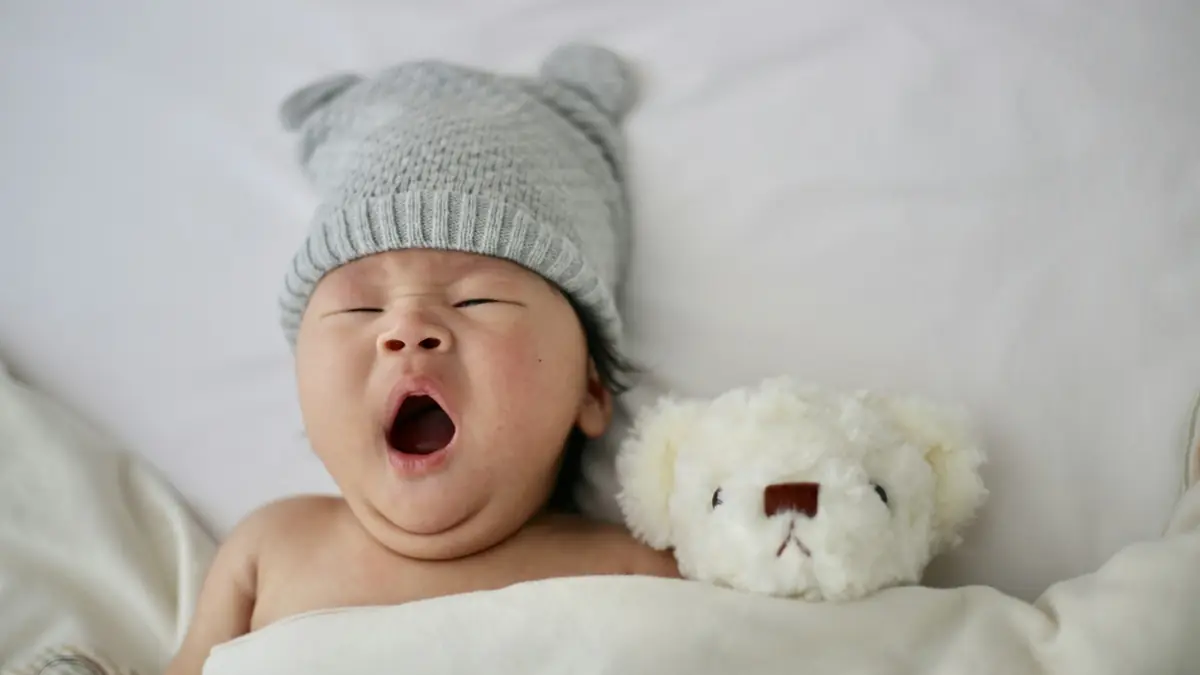 A yawning baby wearing a grey knit hat beside a white teddy bear